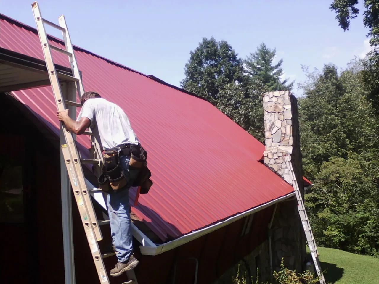 Roofer on a ladder beside a red metal roof and stone chimney — Soffit & Fascia Repair by Roofing Brothers in Seal Beach